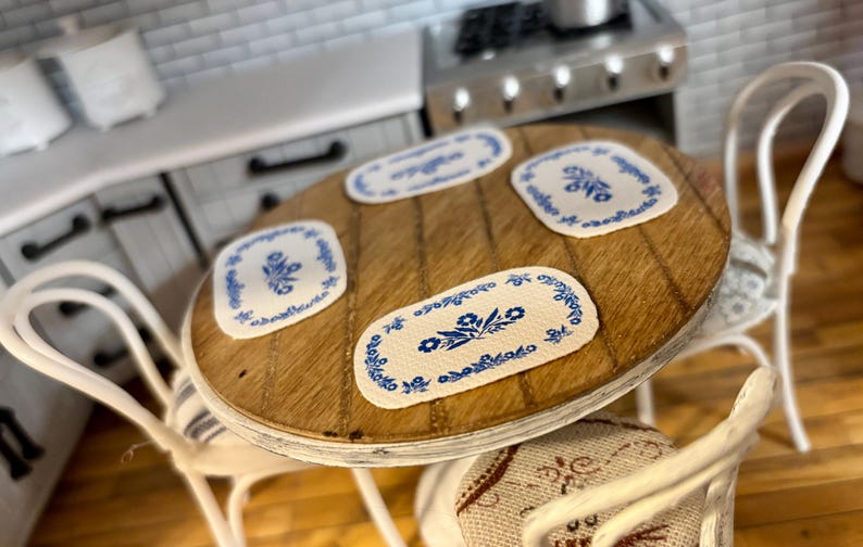 May include: A white wooden table with four white chairs. The table has a wooden top with four blue and white floral placemats.