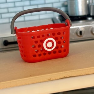 Pu&ograve; includere: Un cesto della spesa in plastica rosso con un manico grigio e un logo Target bianco.