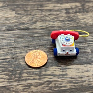 May include: A miniature toy telephone with a red receiver, white body, and blue wheels. The phone has a colorful dial and a smiling face. A penny is next to the toy for size comparison. The toy is on a wooden surface.