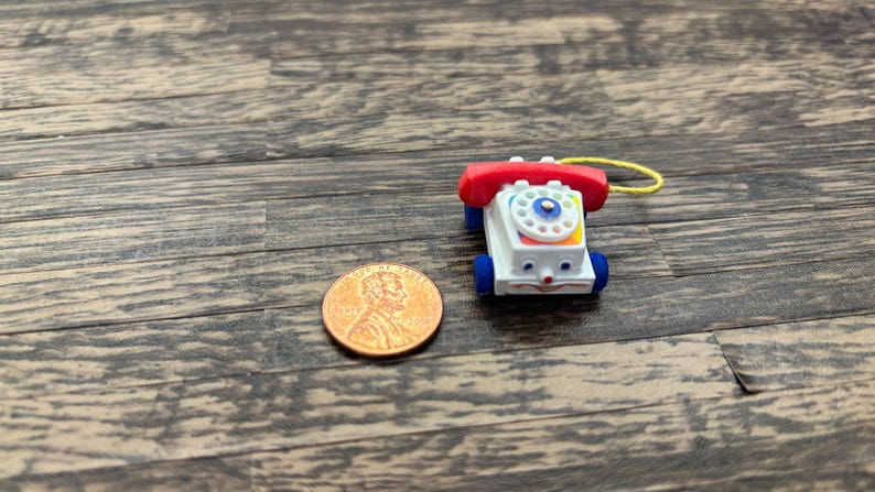 May include: A miniature toy telephone with a red receiver, white body, and blue wheels. The phone has a colorful dial and a smiling face. A penny is next to the toy for size comparison. The toy is on a wooden surface.
