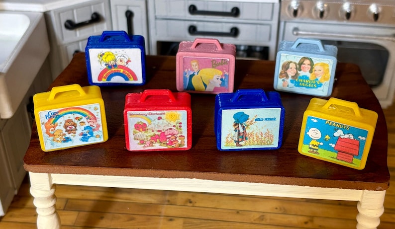 May include: Seven miniature vintage lunchboxes with various cartoon characters. The lunchboxes are yellow, blue, red, and pink. The characters include Care Bears, Strawberry Shortcake, Holly Hobbie, Charlie's Angels, Peanuts, and Barbie.