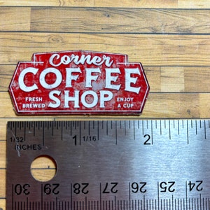 May include: Red and white metal sign with the text "Corner Coffee Shop" in a distressed style. The sign also includes the text "Fresh Brewed" and "Enjoy a Cup".