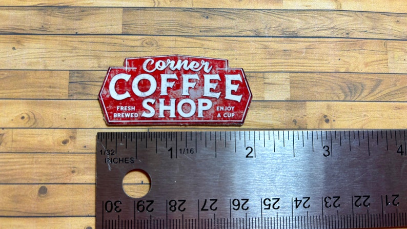 May include: Red and white metal sign with the text "Corner Coffee Shop" in a distressed style. The sign also includes the text "Fresh Brewed" and "Enjoy a Cup".