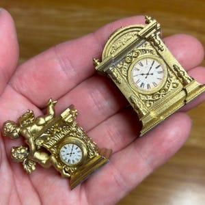 May include: Two miniature gold-coloured clocks. One clock has a rectangular shape with an arched top and Roman numeral clock face. The other clock has a cherub design. Both clocks have intricate detailing and are held in a hand.