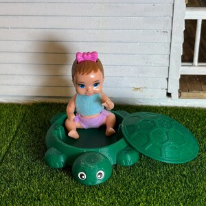 Puede incluir: Un peque&ntilde;o arenero de tortuga verde con tapa. Una mu&ntilde;eca con cabello casta&ntilde;o y un lazo rosa se sienta dentro del arenero.