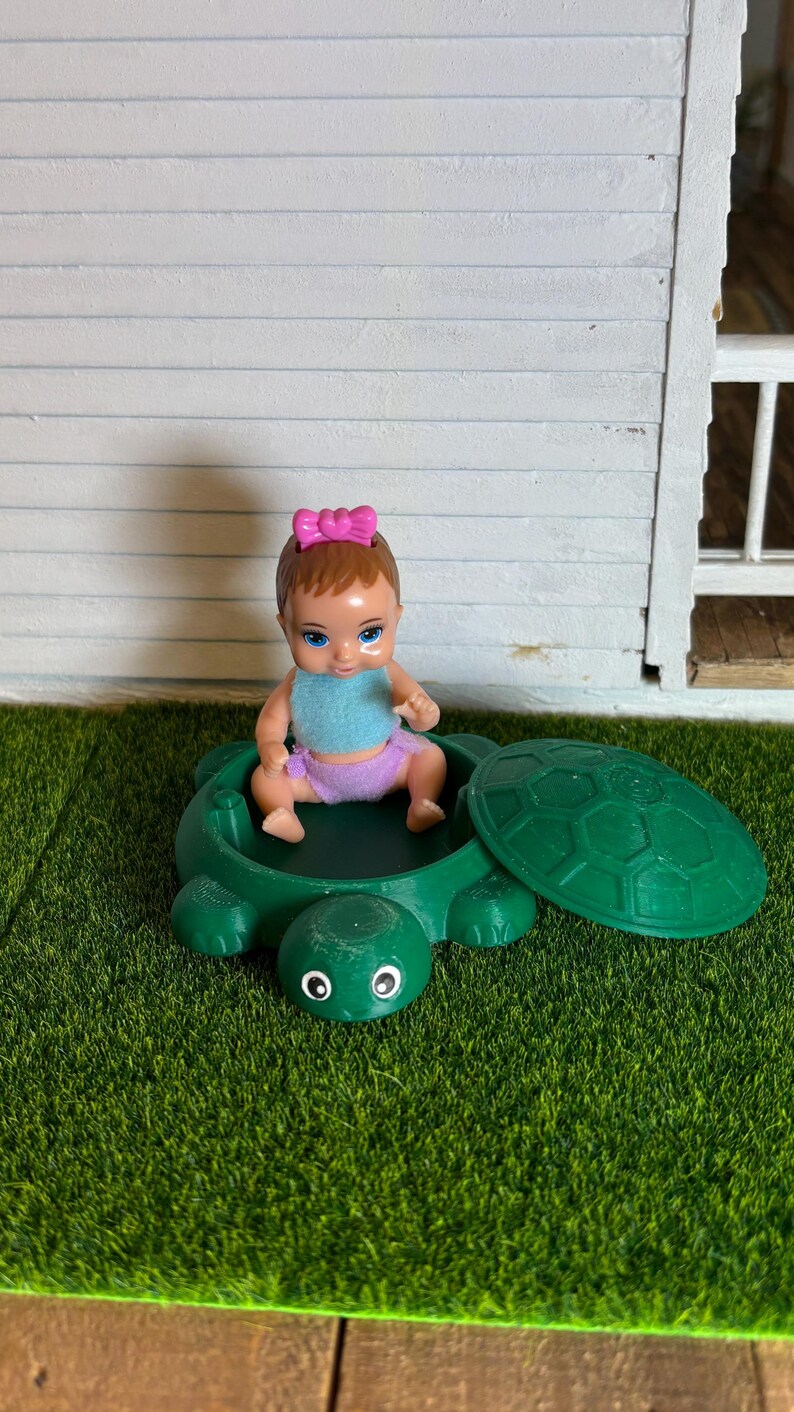 Puede incluir: Un peque&ntilde;o arenero de tortuga verde con tapa. Una mu&ntilde;eca con cabello casta&ntilde;o y un lazo rosa se sienta dentro del arenero.