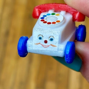 May include: A white toy telephone with a red rotary dial and blue wheels. The toy has a smiling face with blue eyes and a red nose.