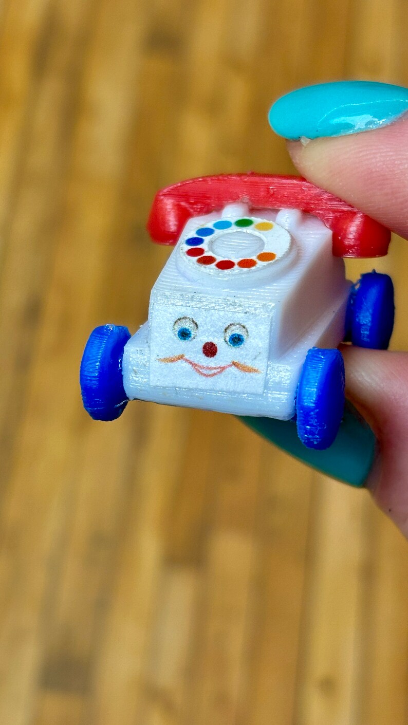 May include: A white toy telephone with a red rotary dial and blue wheels. The toy has a smiling face with blue eyes and a red nose.