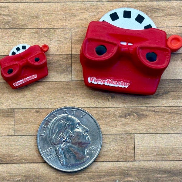 May include: Two red View-Master toys, one large and one small, with black lenses and white film reels. The larger toy has the words "View-Master" printed on it. A quarter is next to the toys for size comparison.