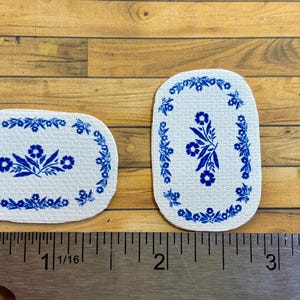 May include: Two white oval-shaped rugs with a blue floral design. The rugs are approximately 1 1/16 inches and 2 inches long.