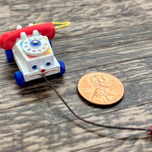 May include: A vintage toy telephone with a red receiver and blue wheels. The white body features a colorful dial and a smiling face. A penny is next to the toy for size comparison.