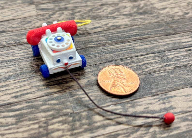 May include: A vintage toy telephone with a red receiver and blue wheels. The white body features a colorful dial and a smiling face. A penny is next to the toy for size comparison.