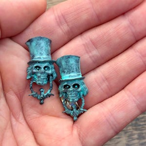 Könnte beinhalten: Zwei türkisgrüne Ohrringe in Schädelform mit Zylinder und Fledermausdetails. Die Ohrringe haben ein verwittertes, antikes Finish, und die Schädel haben leuchtende Augen. Die Ohrringe werden in einer Hand gehalten.