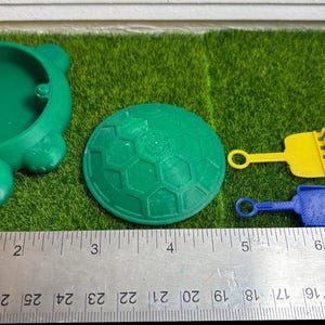 Puede incluir: Un arenero verde en forma de tortuga con una tapa verde, una tapa en forma de panal verde y amarilla, un rastrillo amarillo, una pala azul y un cubo rojo con un asa blanca.