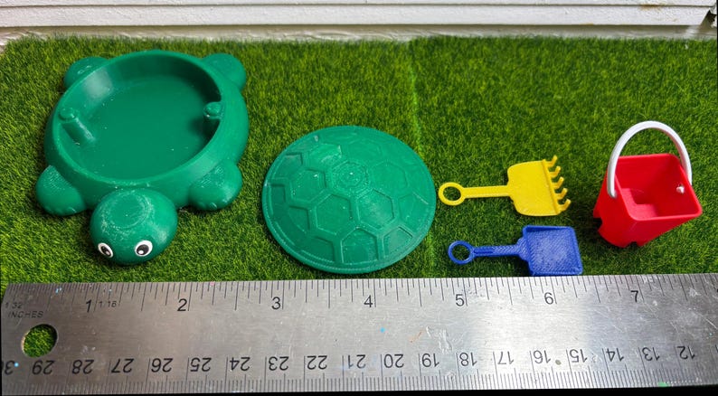 Puede incluir: Un arenero verde en forma de tortuga con una tapa verde, una tapa en forma de panal verde y amarilla, un rastrillo amarillo, una pala azul y un cubo rojo con un asa blanca.