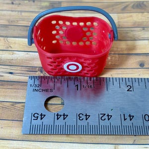 Pu&ograve; includere: Un cestino della spesa in plastica rossa con un manico grigio e il logo Target. Il cestino &egrave; alto circa 2,5 cm e si trova su una superficie di legno.