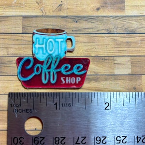 May include: A red and blue metal sign with the words "Hot Coffee Shop" and a blue coffee cup with the word "HOT" on it. The sign is about 1 1/16 inches wide.