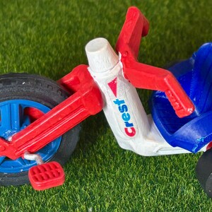 May include: A small tricycle in red, white, and blue, with a black tyre and blue wheel spokes. The tricycle has a seat and handlebars. The word "Crest" is printed on the white body of the tricycle.