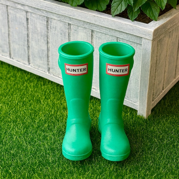 May include: A pair of bright green Hunter rain boots with the brand name displayed on a red and white label. The boots are set on a bed of green artificial grass, with a white planter box and greenery in the background.
