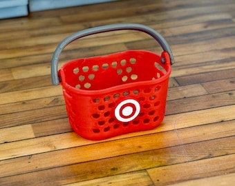 Op de afbeelding: Een rode plastic boodschappenmand met een grijze handvat en een wit Target-logo. De mand is leeg en staat op een houten vloer.