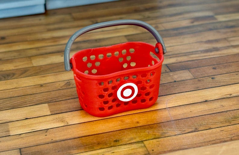 Pu&ograve; includere: Un cesto della spesa in plastica rosso con un manico grigio e un logo Target bianco. Il cesto &egrave; vuoto e si trova su un pavimento di legno.
