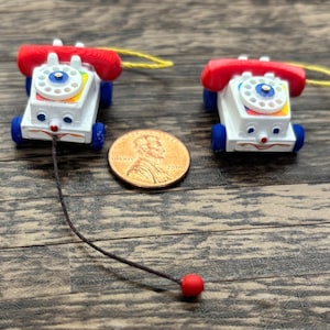 May include: Two miniature toy phones with red handsets, white bodies, and blue wheels. Each phone has a pull string with a red bead. A penny is next to the phones. The phones have a smiling face design.