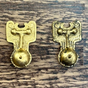 Puede incluir: Dos tiradores de cajón antiguos de color latón con un diseño ornamentado. Cada tirador presenta una parte superior decorativa con un vástago central que conduce a un pomo redondo y texturizado. Los tiradores se muestran sobre una superficie de madera.