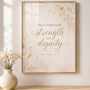 Puede incluir: Una lámina de arte mural rectangular en un marco dorado fino con el texto "She is clothed with strength and dignity" y "PROVERBS 31:25" en letras doradas, decorada con ilustraciones botánicas estilo acuarela en tonos oro y rosa suave.
