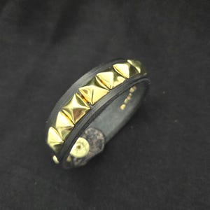 May include: Black leather bracelet with gold pyramid studs. The bracelet features a series of evenly spaced, shiny gold studs along its surface. The bracelet is a dark color, and the studs provide a striking contrast.