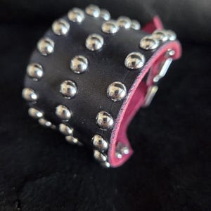May include: A black leather cuff bracelet featuring three rows of polished silver-tone metal studs. The interior of the cuff is lined with a vibrant pink material, and the edges show the pink lining against the black leather exterior.