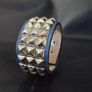 May include: A blue leather cuff bracelet featuring three horizontal rows of silver-toned metal pyramid studs. The interior of the bracelet is a light beige color and includes two silver-toned metal snap fasteners for closure.