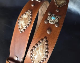 Puede incluir: Un cinturón de cuero marrón con elementos decorativos. El cinturón presenta tachuelas metálicas, detalles de color turquesa y parches en forma de diamante de piel de animal. La hebilla y los herrajes son de color cobre.