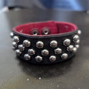 Puede incluir: Una pulsera de cuero negro con tachuelas de metal plateado. El interior está forrado con material rojo. La pulsera se asegura con dos broches metálicos. Este accesorio es adecuado para varios estilos.