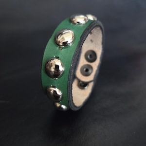 Può includere: Bracciale in pelle verde con borchie color argento. Il bracciale ha una chiusura a scatto. Le borchie sono uniformemente distanziate sulla pelle verde. Sfondo scuro.