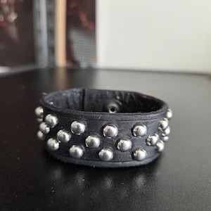 Puede incluir: Pulsera de cuero negro con tachuelas plateadas. La pulsera ancha presenta múltiples filas de tachuelas redondas y metálicas. El cuero parece desgastado, lo que le da un aspecto vintage o atrevido. La pulsera está diseñada para usarse en la muñeca.