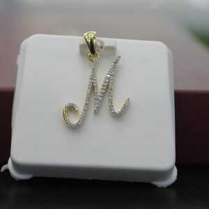 May include: A gold pendant with a cursive letter M set with small diamonds. The pendant is displayed on a white square.
