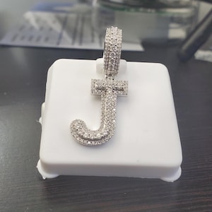 Peut inclure: Pendentif argenté en forme de lettre "J", incrusté de petites pierres scintillantes. Le pendentif est présenté sur un socle blanc. Il comporte une boucle en haut pour une chaîne.