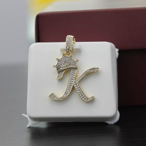May include: Gold pendant featuring the letter K with a crown on top, both encrusted with clear cubic zirconia stones.