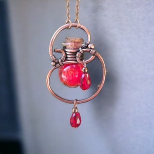 Può includere: Una collana in filo di rame con un ciondolo a forma di fiala di vetro rosso e perline di vetro rosse. La fiala è sospesa in una cornice di filo di rame.