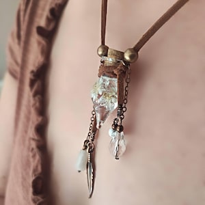 Könnte beinhalten: Halskette mit einem Anhänger aus klarem Glasfläschchen, gefüllt mit getrockneten Blumen. Sie hängt an einem braunen Wildlederband, mit Metallketten, Perlen und einem kleinen Federanhänger. Ein einzigartiges Accessoire.