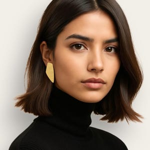 Puede incluir: Una mujer con un jersey de cuello alto negro y pendientes dorados de forma abstracta. Los pendientes tienen una forma plana e irregular. La mujer tiene el pelo oscuro y los ojos marrones y mira directamente a la cámara.