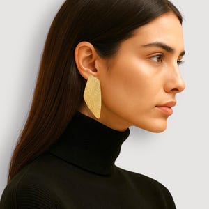 May include: A gold-colored, abstract-shaped earring is worn on the ear of a person wearing a black turtleneck. The earring has a textured surface and a unique, asymmetrical design. The background is a neutral, off-white color.