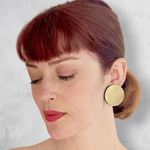 Puede incluir: Una mujer con cabello rojo lleva un par de pendientes redondos de oro. Los pendientes son grandes y tienen una superficie texturizada.