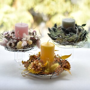 Könnte beinhalten: Drei Kerzenhalter mit Trockenblumenkränzen. Die Kerzen sind rosa, gelb und grau. Die Kränze sind aus Zweigen und Trockenblumen gefertigt.