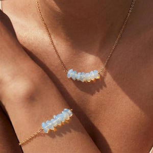 Könnte beinhalten: Ein goldfarbenes Halsketten- und Armband-Set. Jedes Schmuckstück hat eine Reihe kleiner, hellblau-weißer Opalsteine. Der Schmuck ist zart und scheint auf der Haut einer Person getragen zu werden.