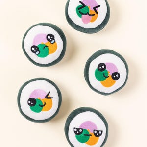 Puede incluir: Cinco rollos de sushi de peluche con caras bordadas. Cada rollo presenta un centro de arroz blanco, una envoltura de algas verdes y rellenos coloridos de naranja, verde y rosa. Los rollos tienen varias expresiones faciales, incluyendo guiños, sonrisas y gafas de sol.