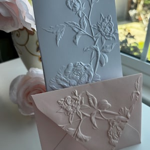 May include: Two embossed cards with floral designs. The top card is light blue with white flowers and leaves. The bottom card is pink with white flowers and leaves.