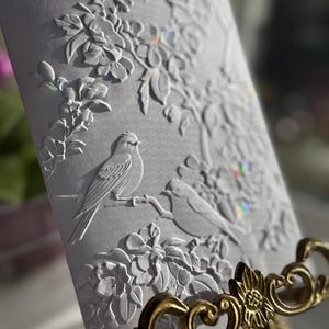 May include: A white embossed card with a floral design and two birds. The card is on a gold stand.