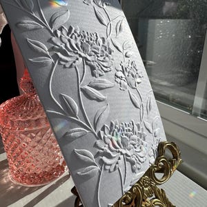 May include: White card with an embossed floral design. The card is on a gold metal stand. The card has a raised design of flowers and leaves.