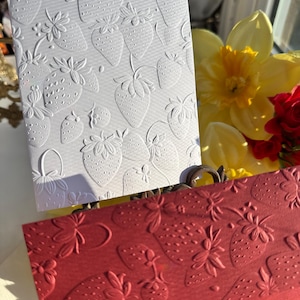 May include: Two embossed cards, one white and one red, feature a repeating strawberry pattern. The white card has a textured surface with detailed strawberry and floral designs. The red card displays a similar strawberry pattern. The cards are part of a stationery set.